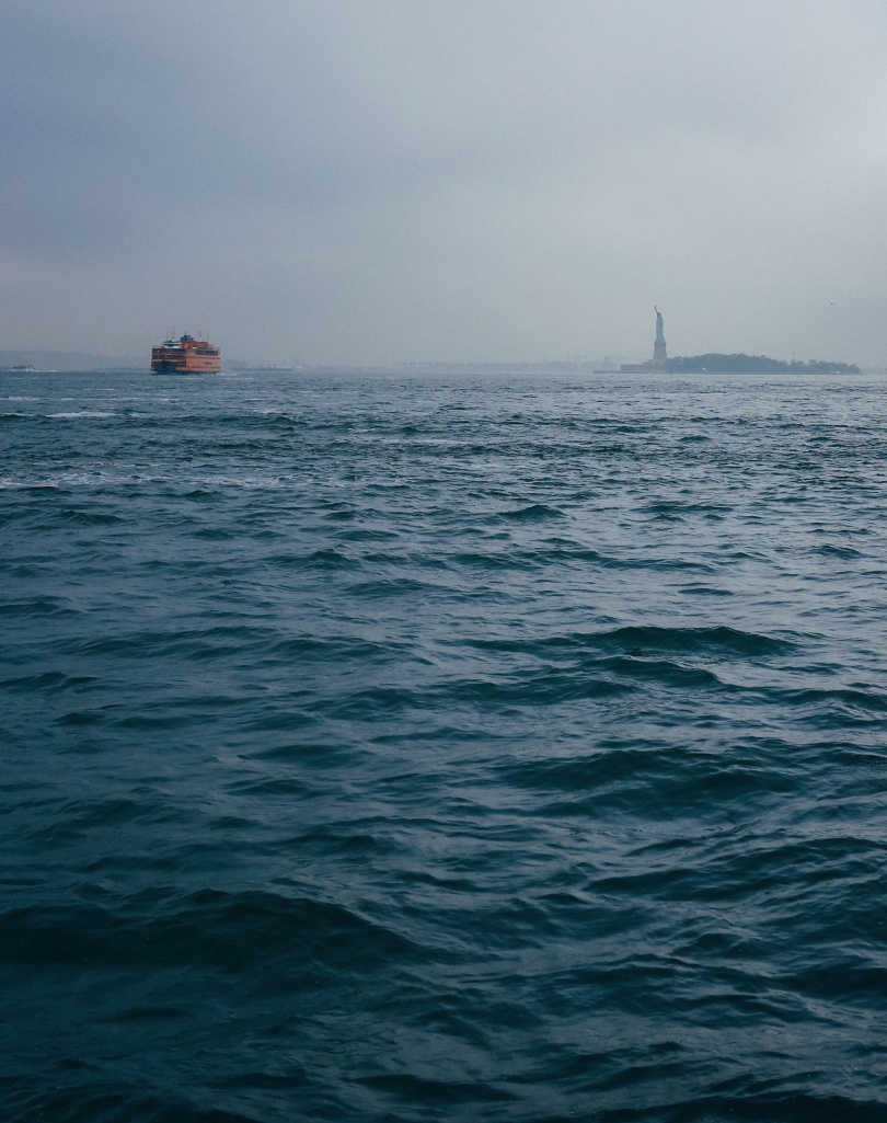 The ocean on a cloudy day. In the distance you can see a ferry and the Statue of Liberty.